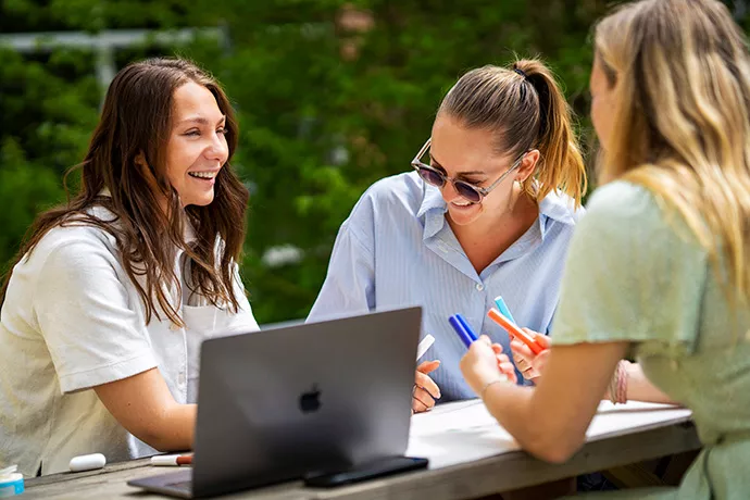 3 happy student sitting around an outdoor table with an open laptop. Photo