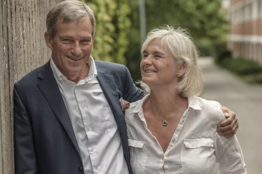 A man in his 60s holds his arm around a woman, his wife. She looks at him and smiles. Photo.